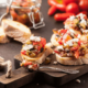 Tranches de pain grillé garnies de légumes rôtis, de tomates fraîches et de fromage feta, présentées sur une planche en bois avec un pot de conserve en arrière-plan, symbolisant une mise en bouche gourmande.